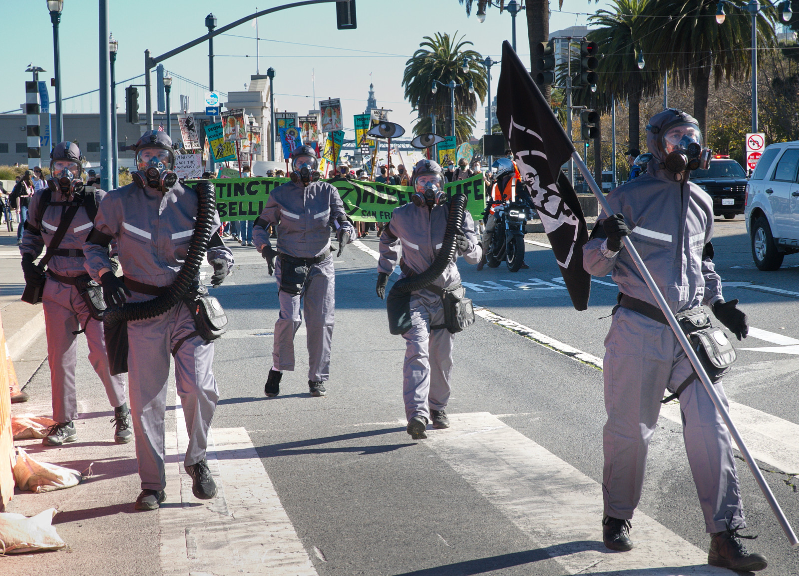 Five characters walk down the street in San Francisco, wearing masks and grey hazmat suits